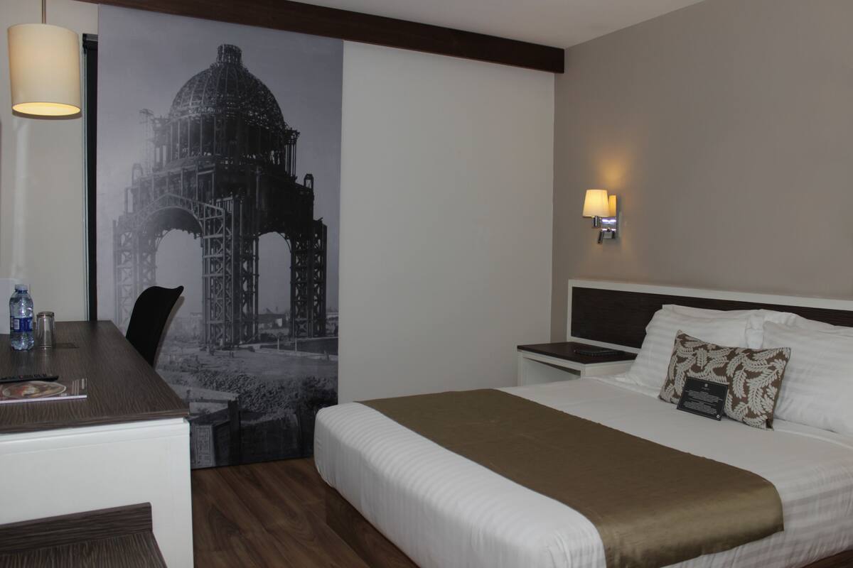 A hotel room with a bed, a desk, a chair, a lamp, and a wall-mounted light fixture.
