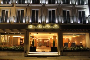 L’extérieur de l’hôtel Plaza Revolución, avec son entrée bien éclairée, ses grandes fenêtres et une enseigne au-dessus de l’entrée.