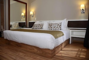 Une chambre d’hôtel avec un grand lit, une tête de lit en bois, deux tables de chevet, une commode, un miroir et des lampes fixées au mur.