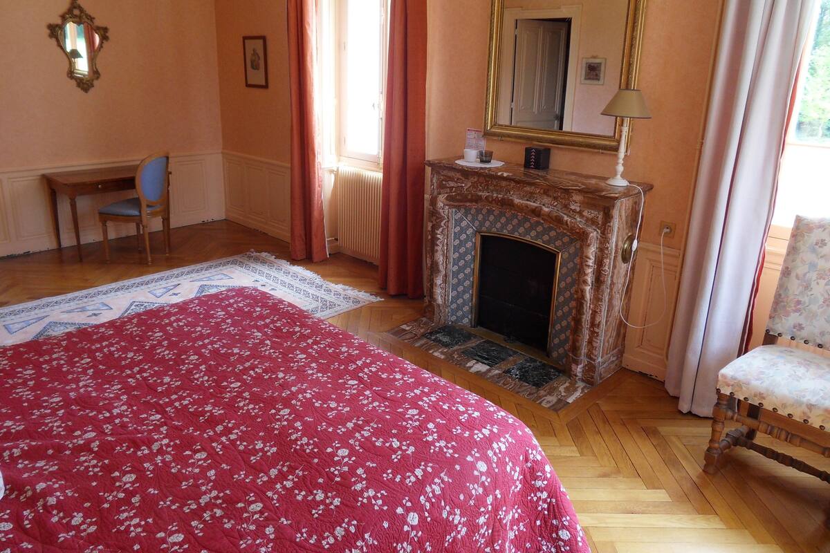A bedroom with a fireplace, a bed with a floral bedspread, a chair, and a mirror on the wall.