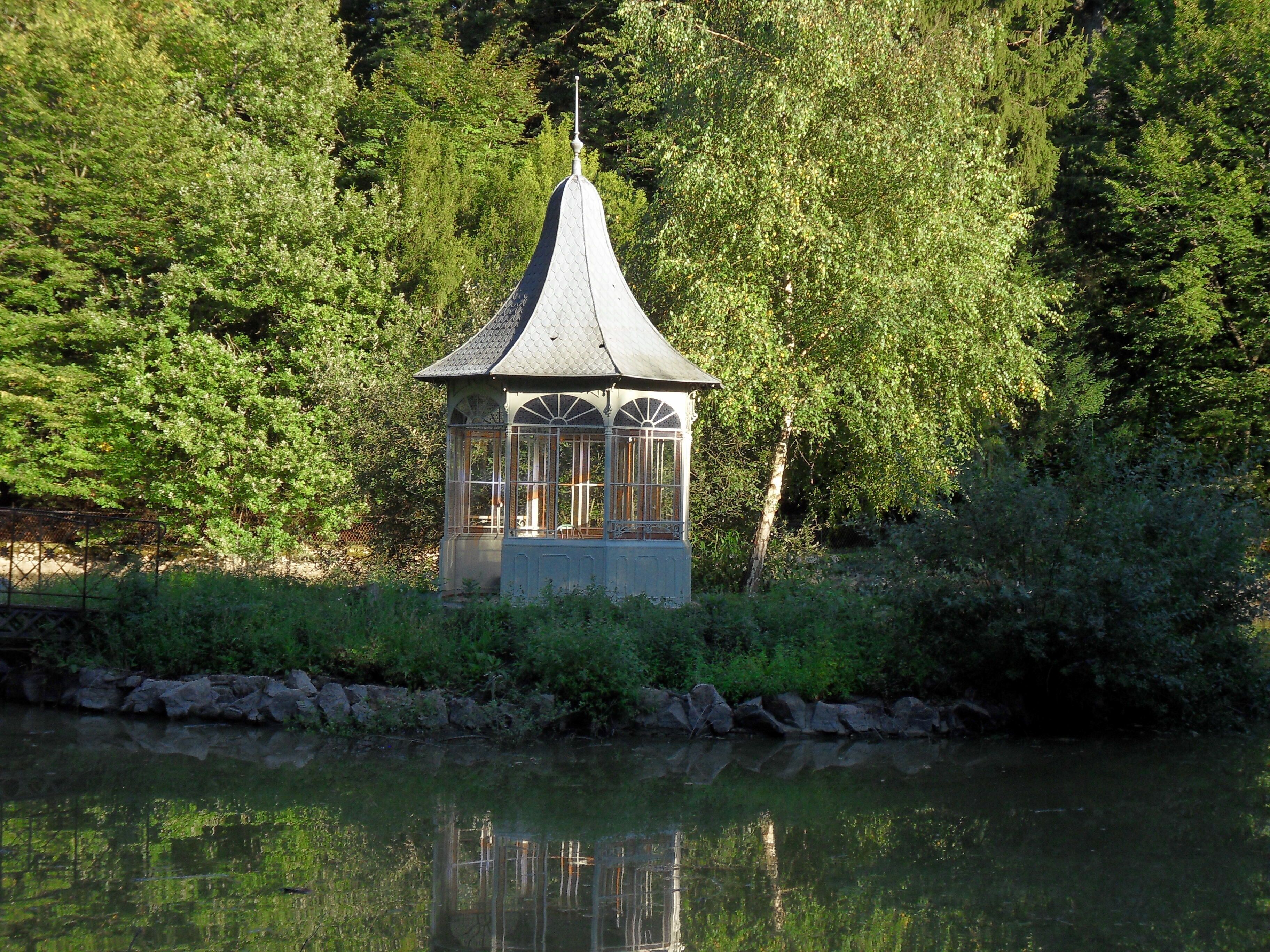 A gazebo with a pointed roof and glass panels, surrounded by dense greenery and a calm body of water.