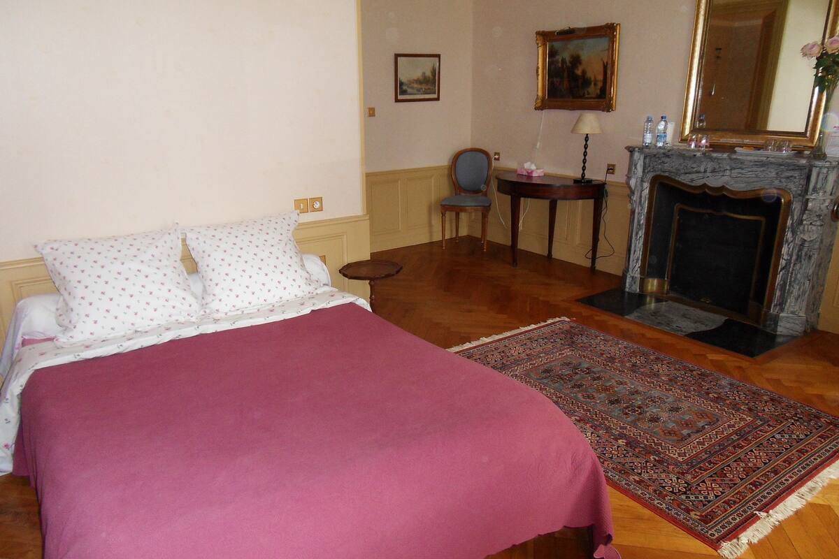 A bedroom with a bed, a chair, a small table, a fireplace, and a rug.