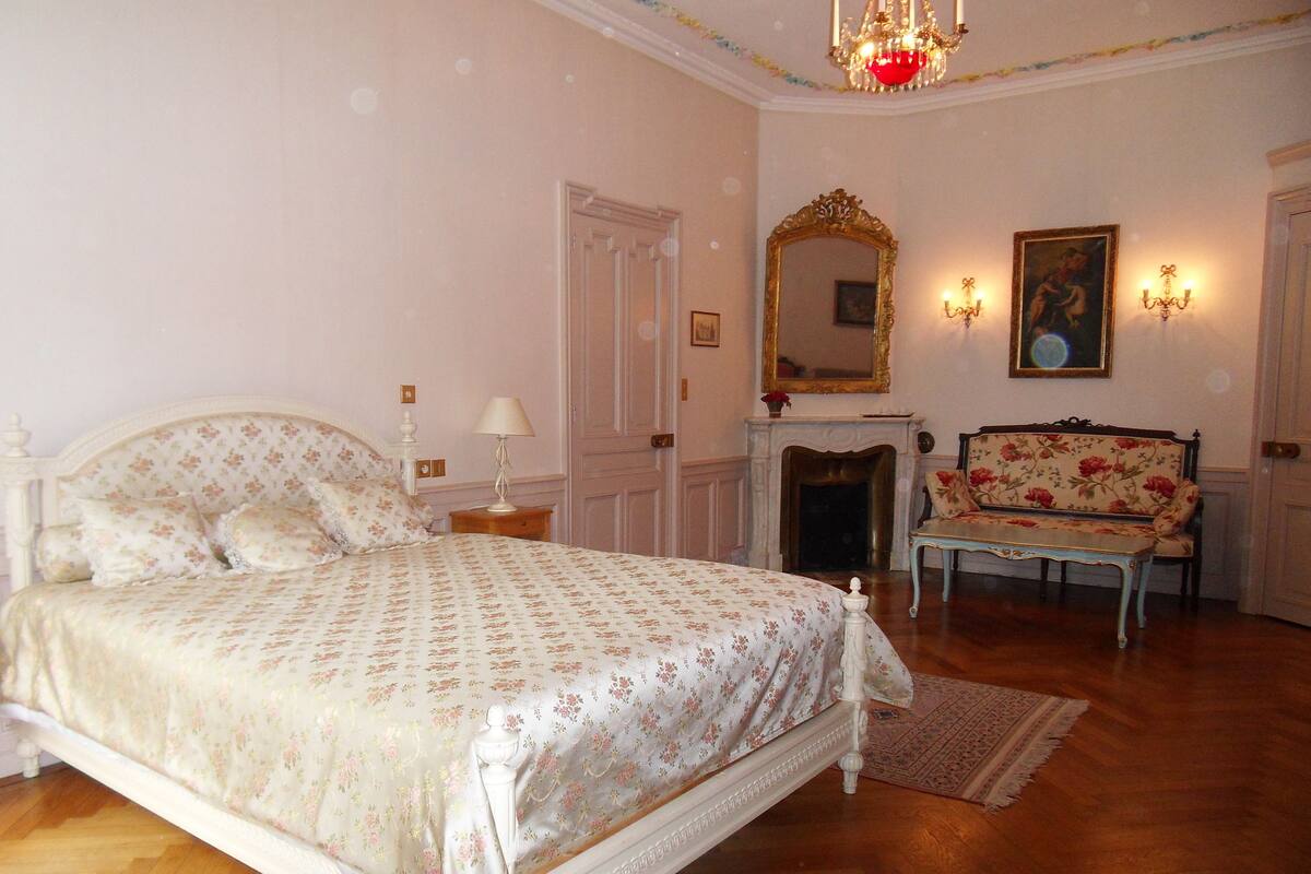 A bedroom with a large bed, a fireplace, a bench, and a mirror.