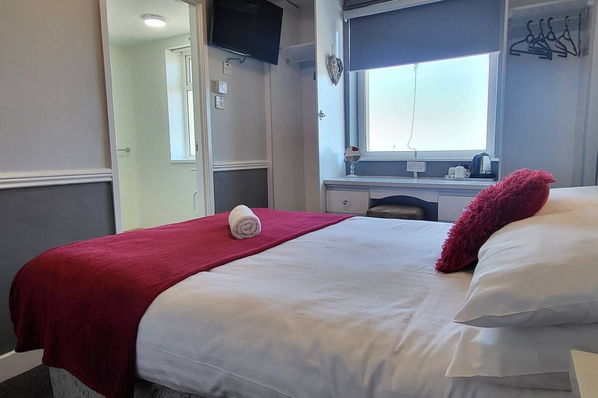 Hotelkamer met een bed, een rode plaids, een wit kussen, een rode deken, een klein tafeltje met een waterkoker en een raam met uitzicht.