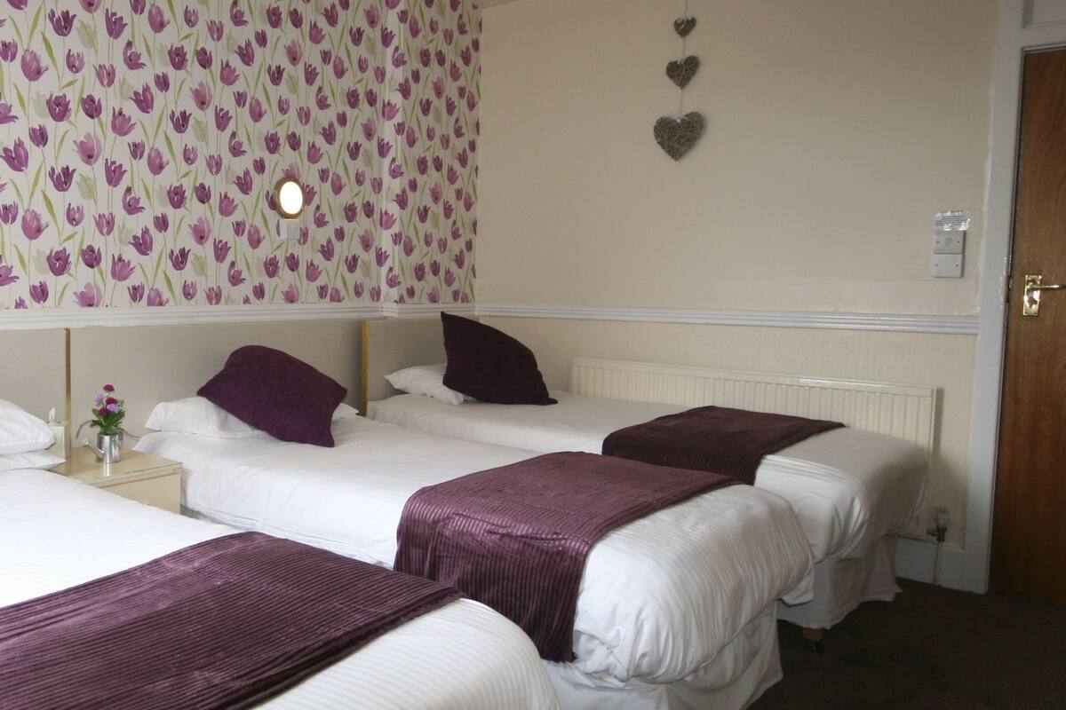 Een hotelkamer met vier eenpersoonsbedden, elk voorzien van een paarse deken en kussen. De kamer heeft behang met bloemenmotieven en een klein nachtkastje met een bloemenvaas.
