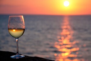 Una copa de vino blanco sobre una roca, con una puesta de sol y el océano al fondo.