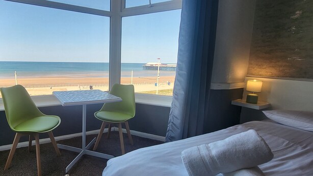 A hotel room with a large window overlooking a beach, a bed, a small table, and two chairs. - Blenheim Mount Hotel (Blackpool)