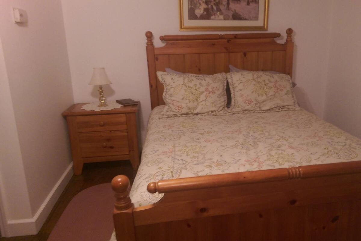 A wooden bed with a floral bedspread and two pillows, a nightstand with a lamp and a book, and a framed picture on the wall.