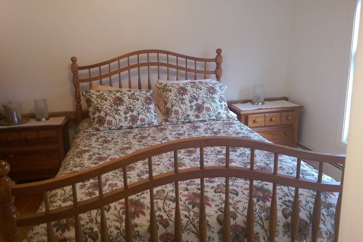 A bedroom with a wooden bed frame, floral bedding, a nightstand with a lamp, and a dresser with drawers.