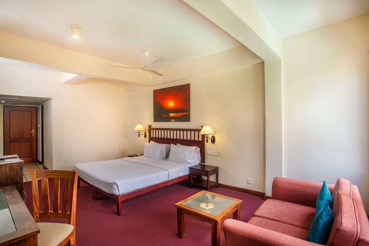 A hotel room with a bed, a desk, a chair, a sofa, a ceiling fan, and a painting on the wall.