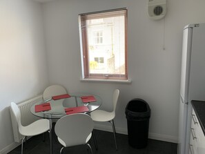 Una habitación pequeña y moderna con una mesa redonda de vidrio, sillas blancas, una ventana con persianas, un radiador, un cesto de basura negro y un refrigerador blanco.