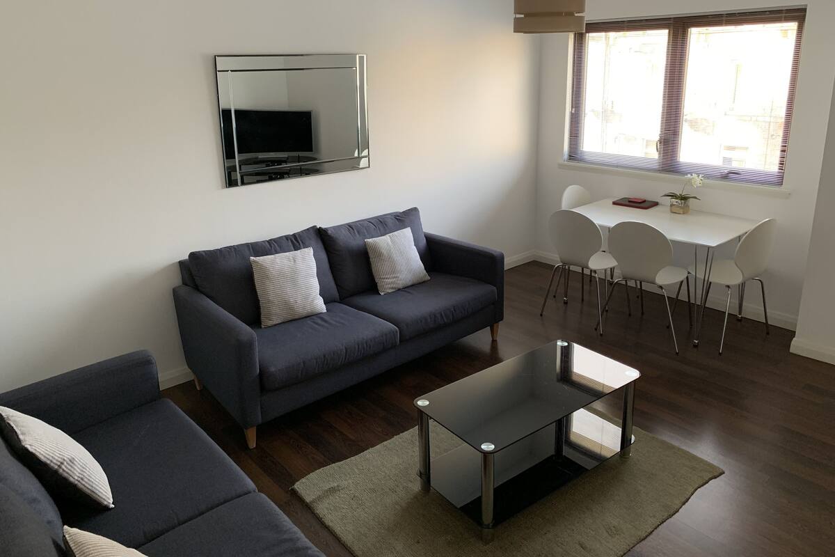 A modern living room with a grey sofa, a glass coffee table, and a dining area with white chairs.