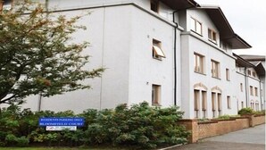 Un immeuble à plusieurs étages, doté d’un toit plat et de fenêtres rectangulaires. Un panneau est placé au sol pour indiquer les restrictions de stationnement.