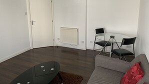 Un salon moderne avec un canapé gris, une petite table ronde, deux chaises noires, une porte blanche et un radiateur blanc.