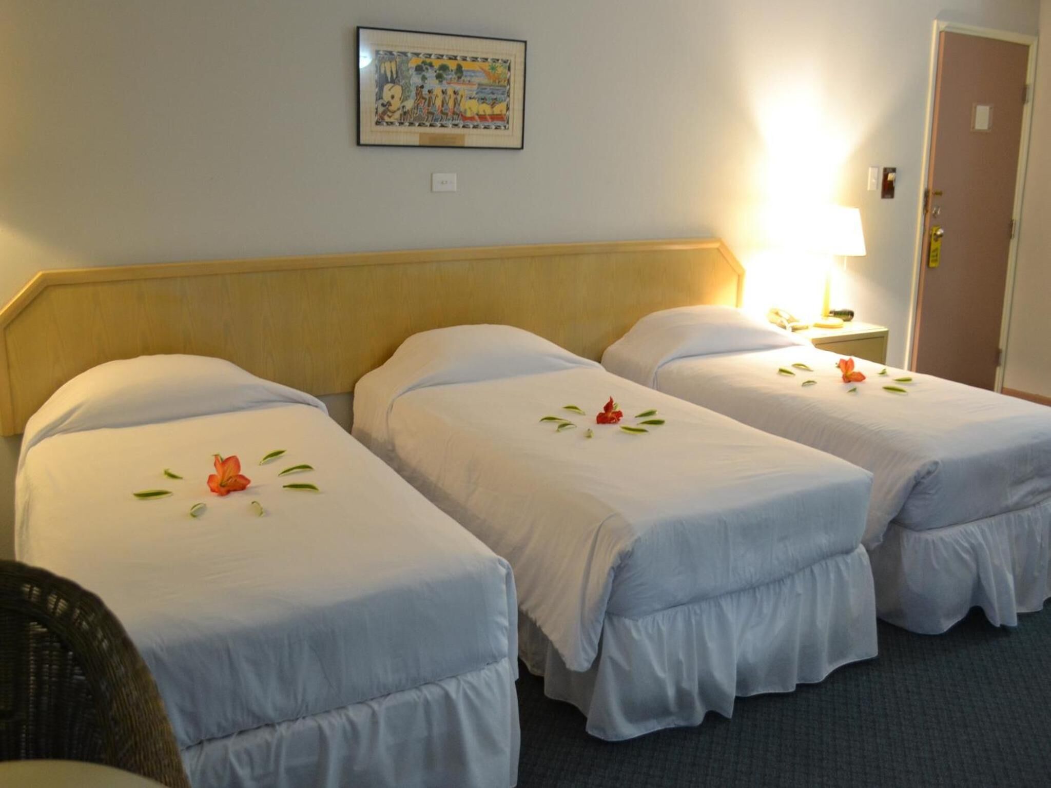 Une chambre d’hôtel avec trois lits simples, chacun recouvert d’une courtepointe blanche et agrémenté d’un petit bouquet de fleurs. Un tableau encadré est accroché au mur au-dessus des lits, et une table de chevet avec une lampe se trouve à droite.