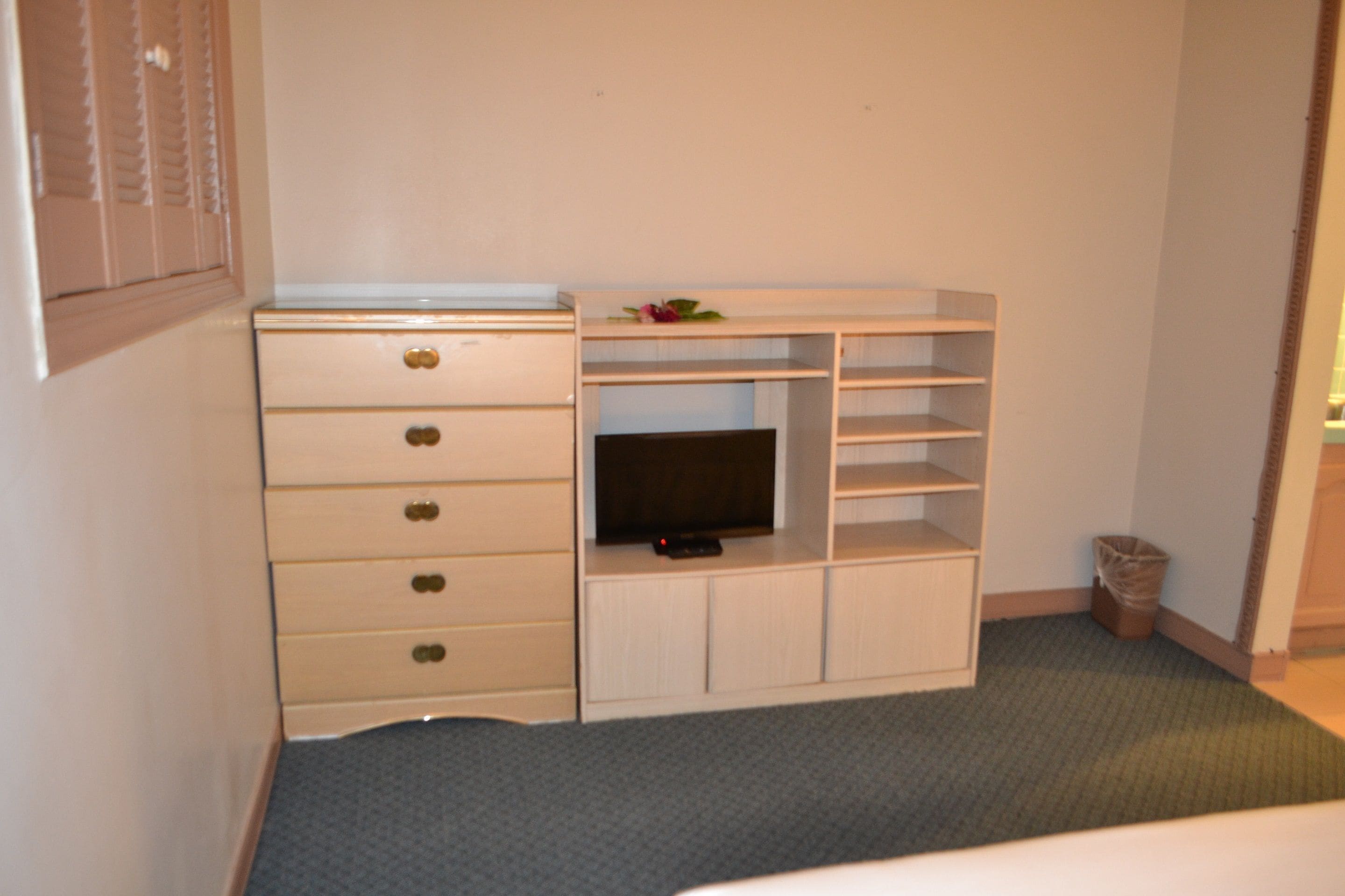 A small room with a white dresser, a television on a shelf, and a trash can.