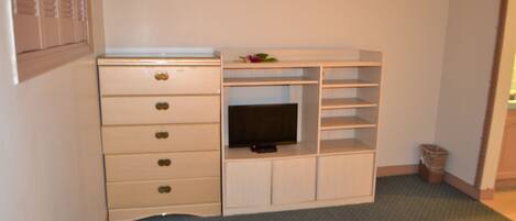 A small room with a white dresser, a television on a shelf, and a trash can.