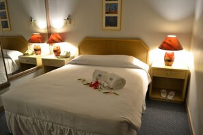 Ein Hotelzimmer mit einem Bett, zwei Nachttischen mit Lampen, einem Spiegel und einem kleinen Regal mit Hausschuhen.