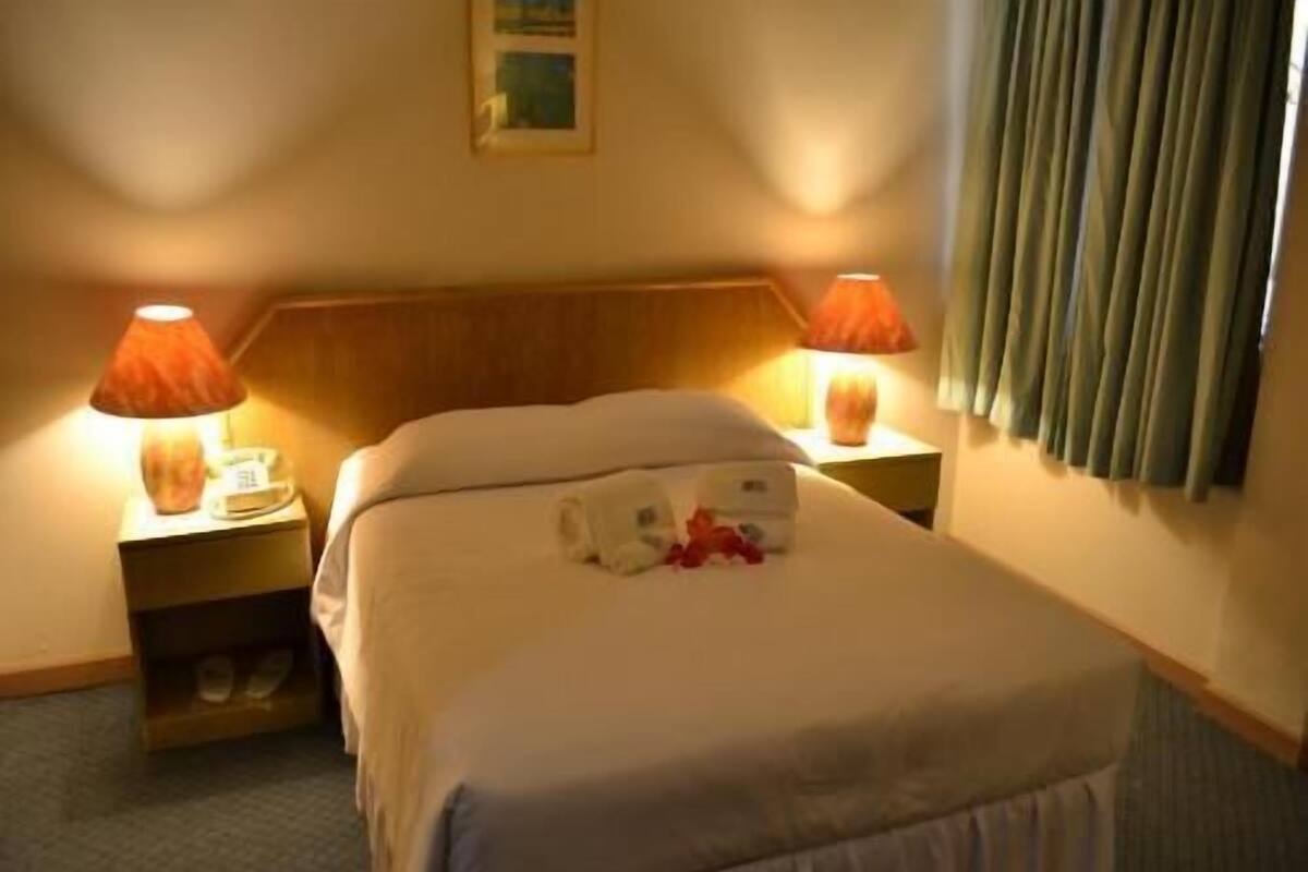 Una habitación de hotel con una cama, dos lámparas de noche, una mesita de noche y un ventanal con cortinas.