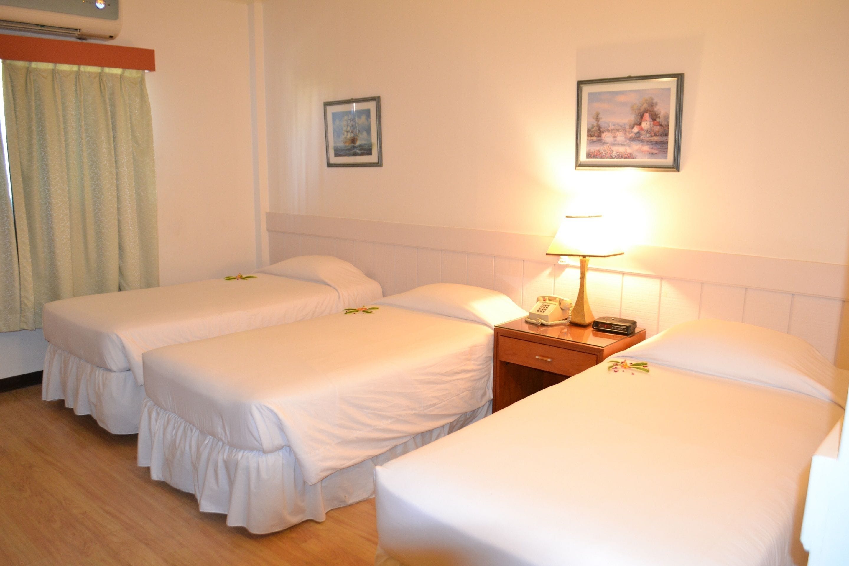 Habitación de hotel con tres camas individuales, una mesita de noche con lámpara y dos cuadros enmarcados en la pared.