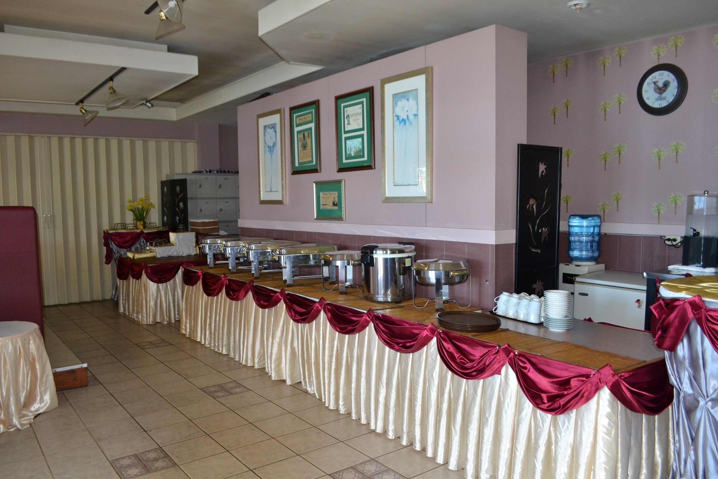 A banquet hall with a long table covered in a white tablecloth with red sashes, surrounded by various food service items.