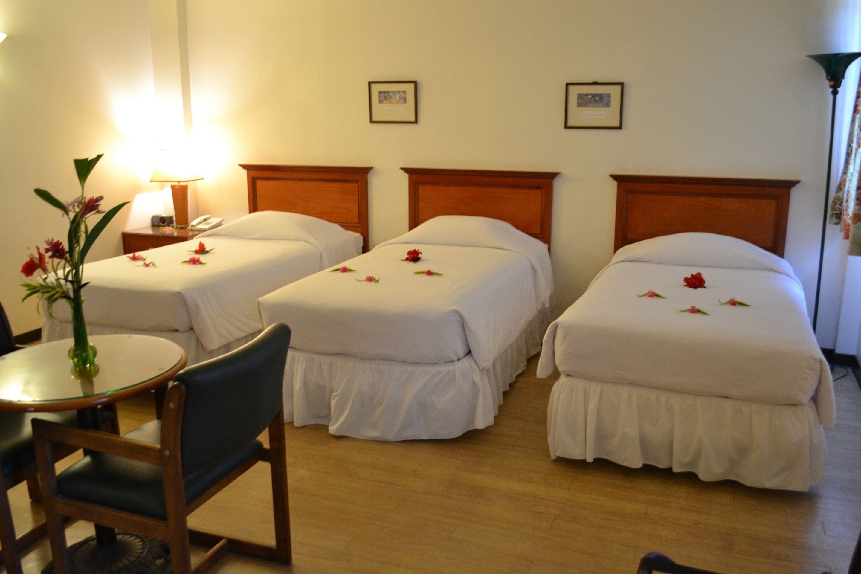 A hotel room with two single beds, a small round table with a chair, a lamp, and a vase with flowers.
