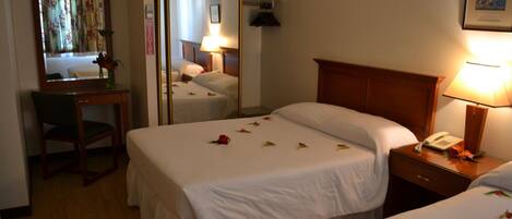 A hotel room with two beds, a desk, a chair, a mirror, and a bedside table with a lamp.
