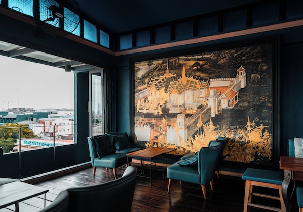 A modern interior with a large mural of a traditional building, seating, and a view of the city. - Baan Wanglang Riverside (Bangkok)