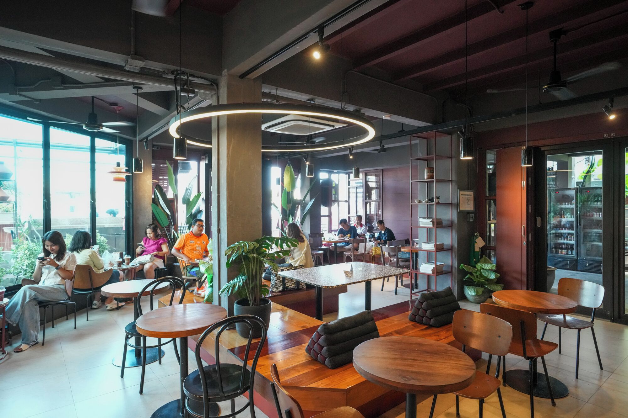 A modern cafe with round tables and chairs, a central seating area with a curved light fixture, and a view of the street outside.