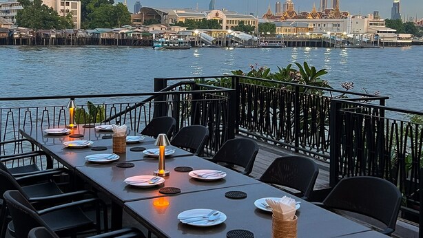 Un área de comedor al aire libre con mesas preparadas para dos, con vista a un frente marítimo con edificios al fondo.