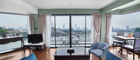 A spacious living room with a large window overlooking a cityscape, a flat-screen TV, a dining area, and a sofa with colorful cushions.