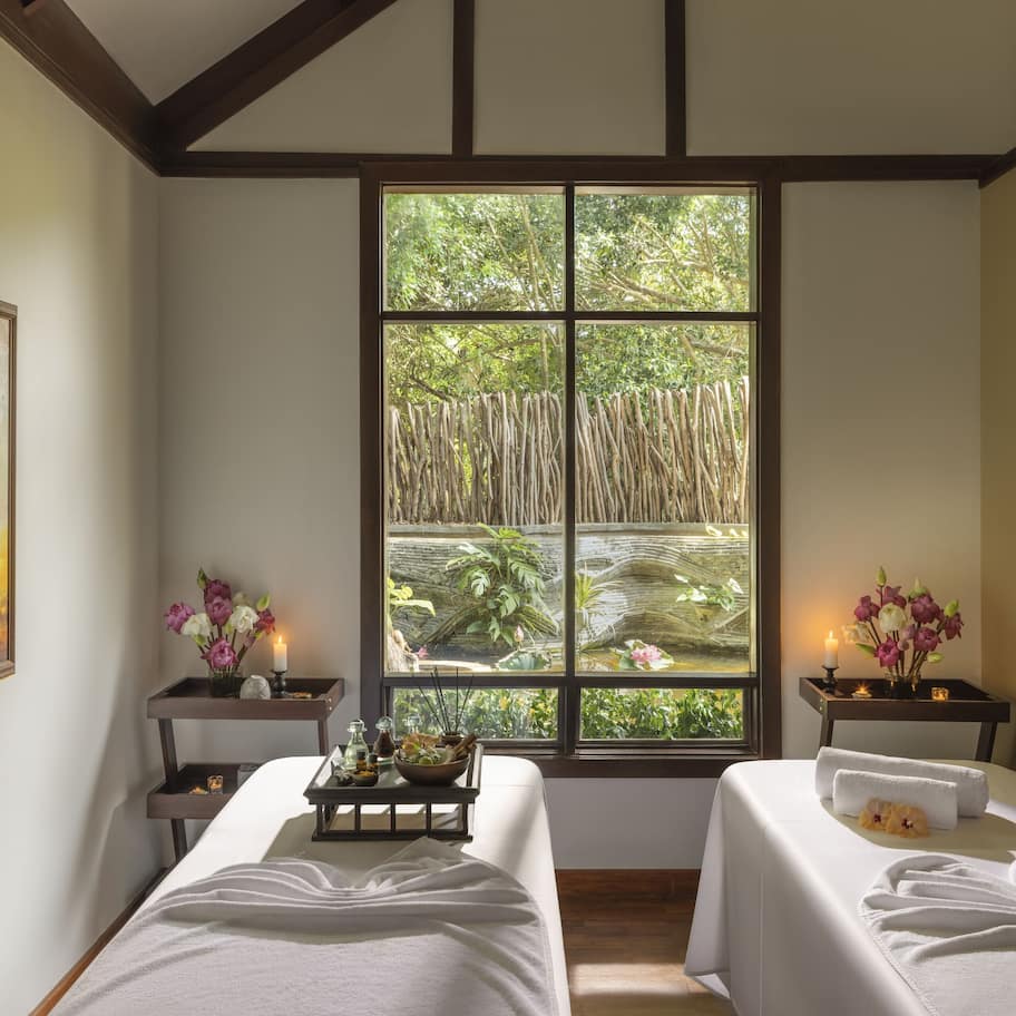 A room with two beds, a large window with a view of greenery, a painting on the wall, and a tray with toiletries.