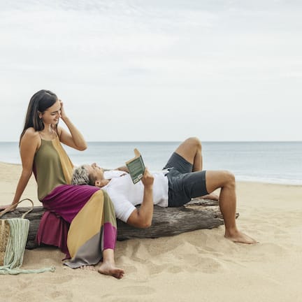 A couple relaxing on a log on a sandy beach with the ocean in the background.