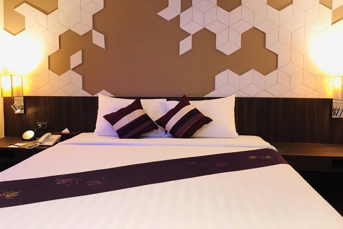 A hotel room with a geometric wall design, a neatly made bed with two pillows, a bedside table with a lamp, and a phone.