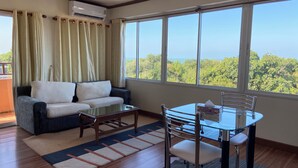 Una sala de estar con sofá, mesa de centro, mesa de comedor con sillas y ventanas grandes que ofrecen vistas a zonas verdes.