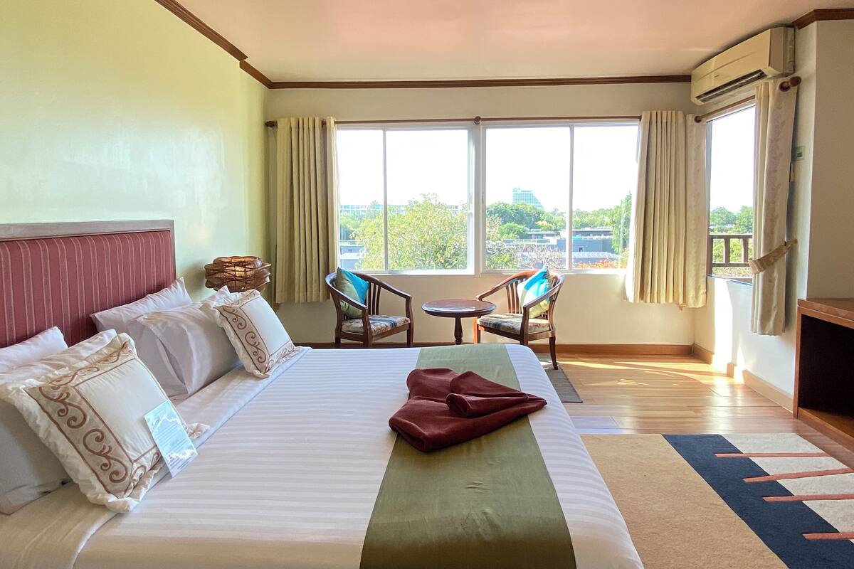 Quarto de hotel com uma cama grande, duas cadeiras, uma mesinha e vista para a cidade através de grandes janelas.
