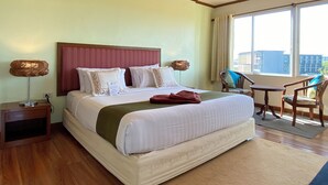 Habitación de hotel con una cama grande, cabecera de madera, mesita de noche con lámpara, una silla y vistas a edificios desde la ventana.