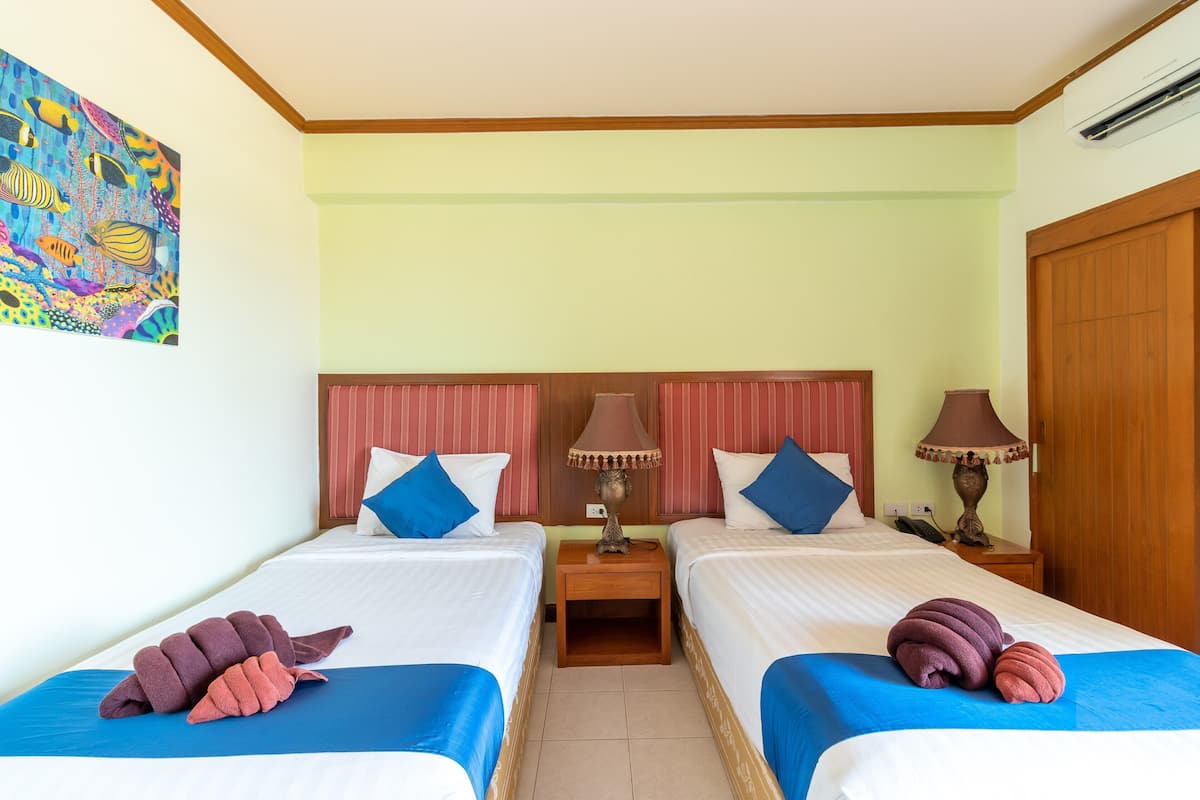 Quarto de hotel com duas camas, uma mesinha de cabeceira com luminárias, um quadro na parede e uma porta de madeira.