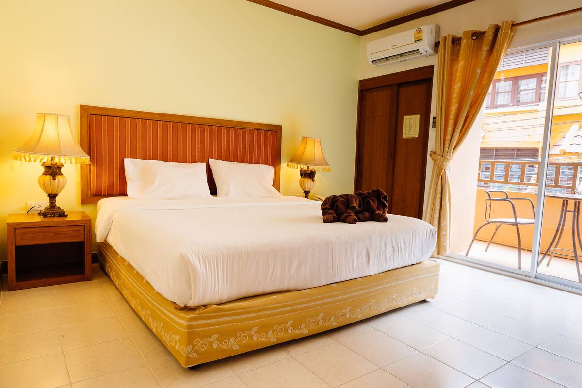 Quarto de hotel com cama grande, duas luminárias de cabeceira, cabeceira de madeira, janela com cortinas e varanda com uma cadeira.