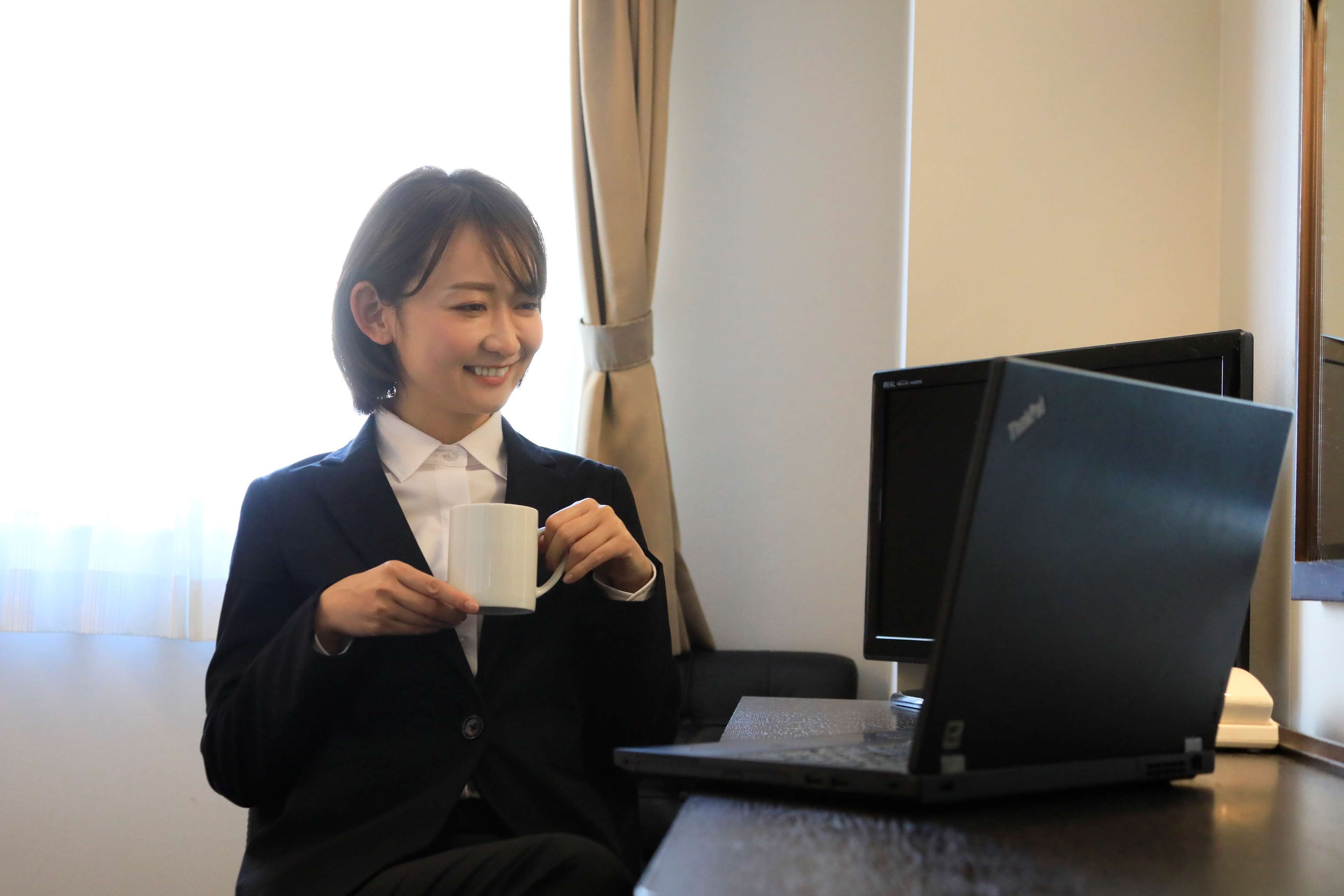 Une personne vêtue d’un costume noir est assise à un bureau, devant un ordinateur portable, et tient une tasse blanche.