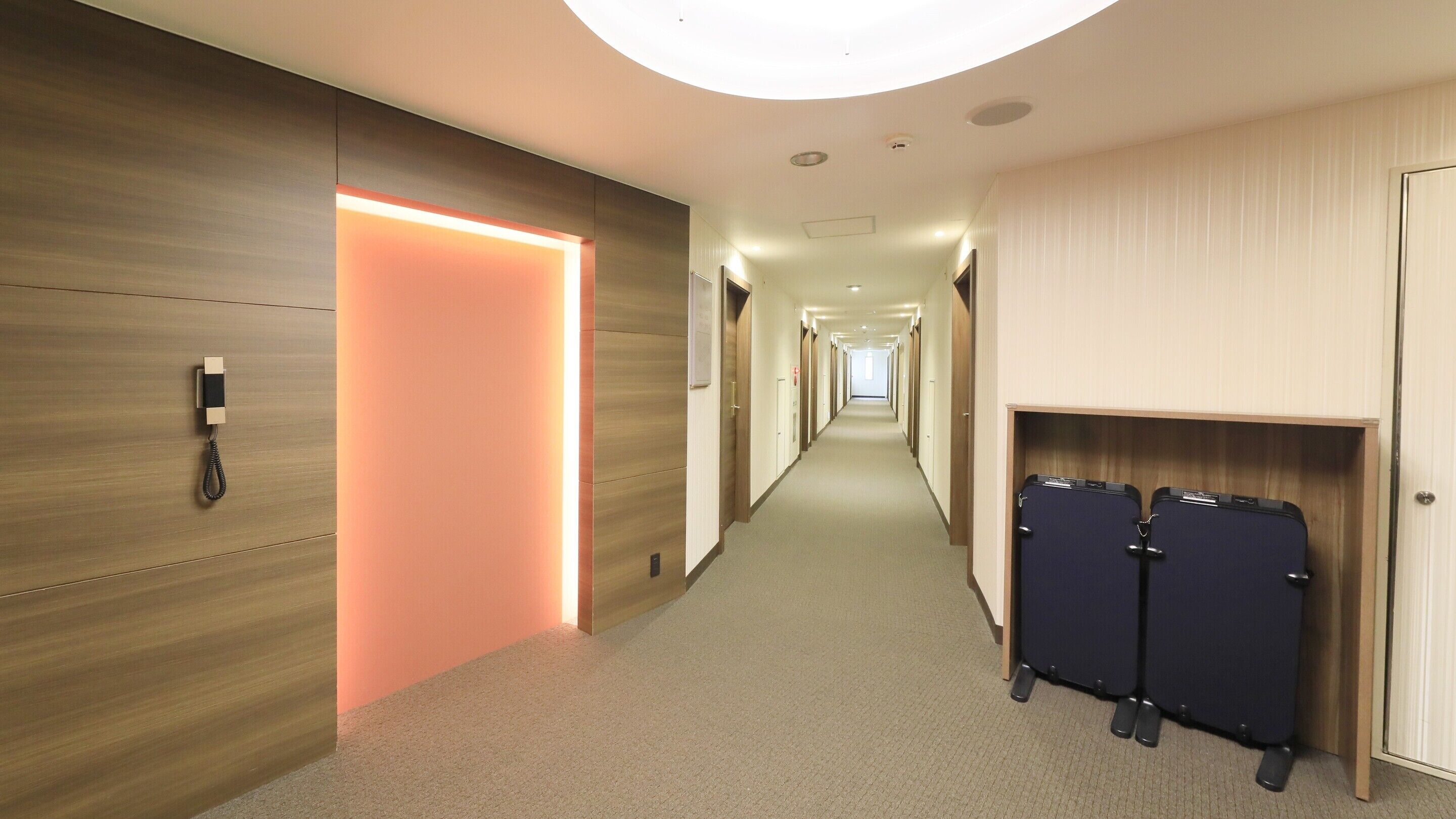 A hotel corridor with a wooden wall on the left, a lit door at the end, and a luggage storage area on the right.