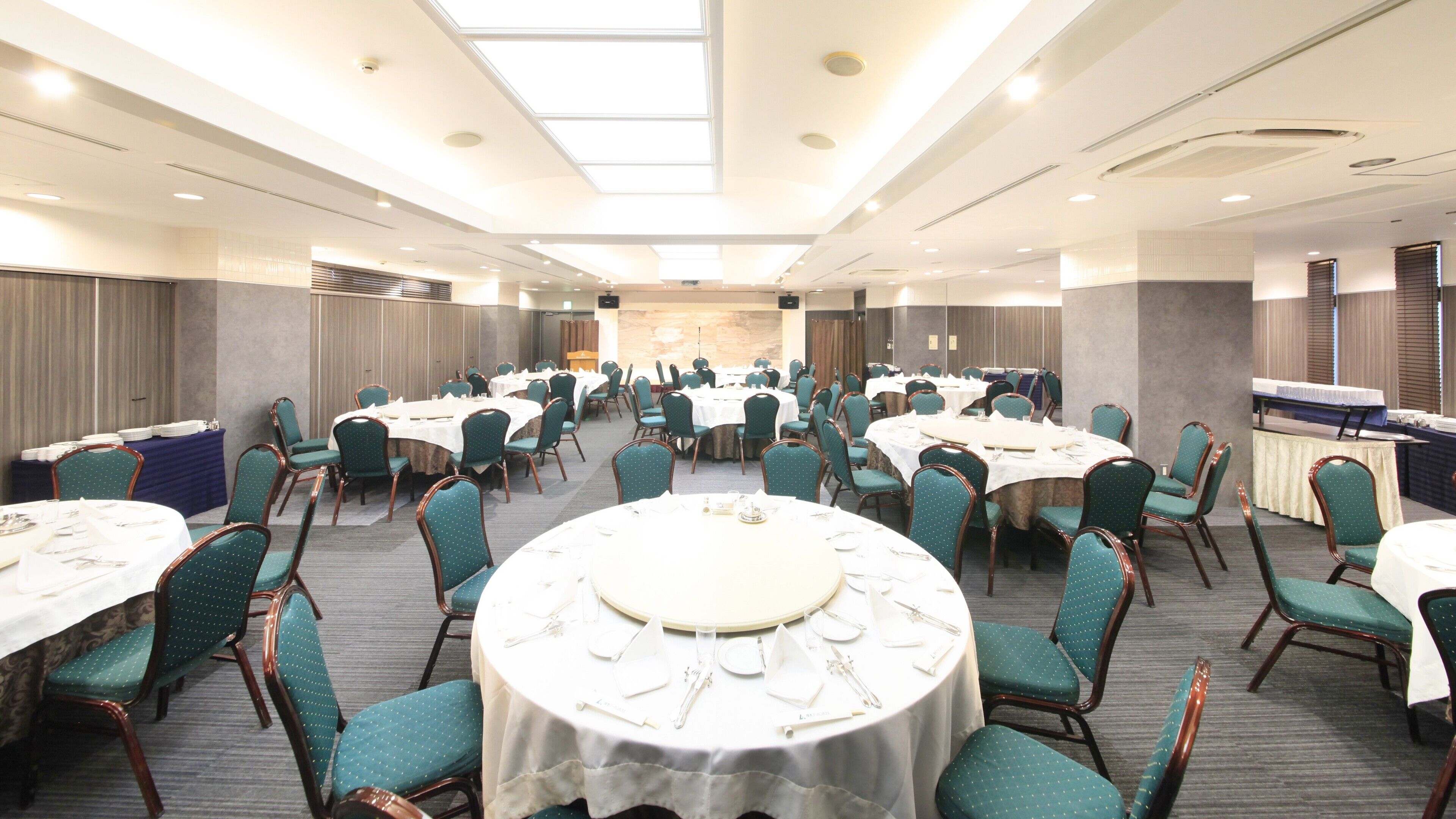 Un salón de banquetes con mesas redondas y sillas, un escenario y una larga barra.