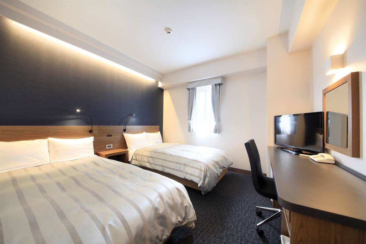 Ein Hotelzimmer mit zwei Betten, einem Schreibtisch mit Fernseher und einem Fenster mit Vorhängen.
