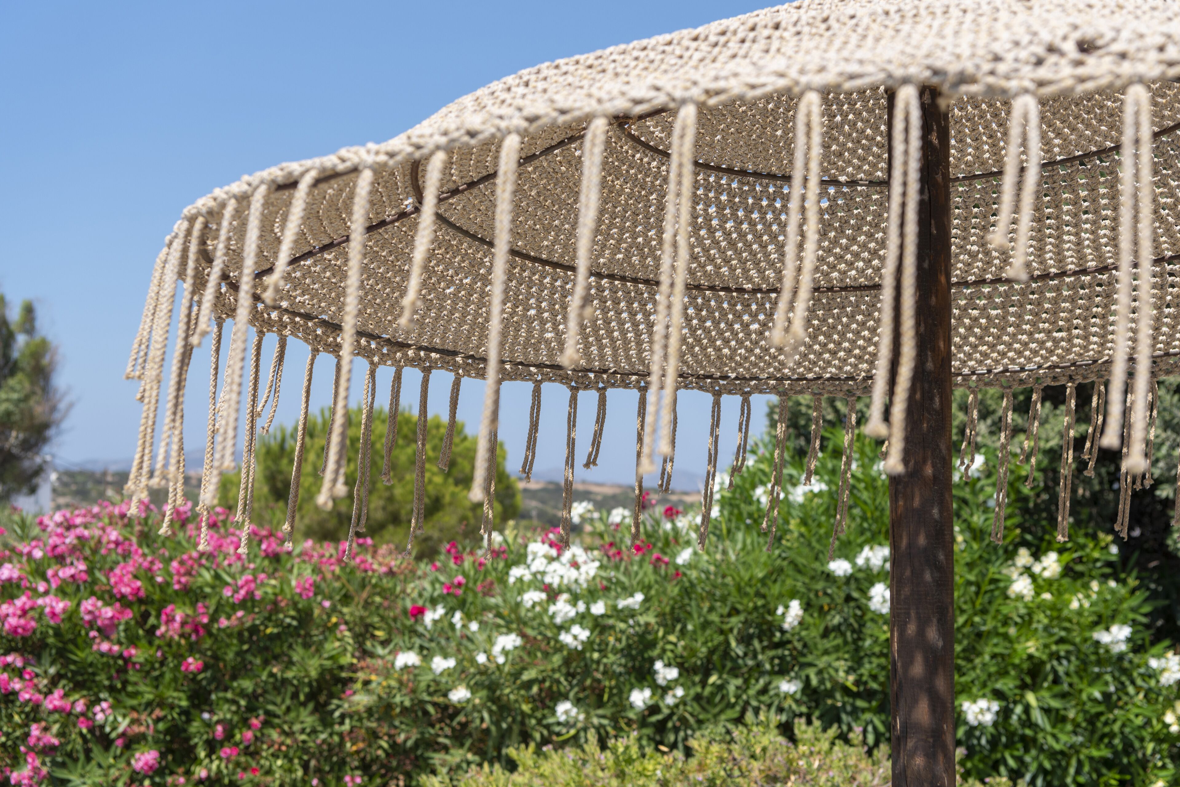 A large, woven outdoor structure with ropes hanging from it, set against a backdrop of flowering bushes and a clear sky.