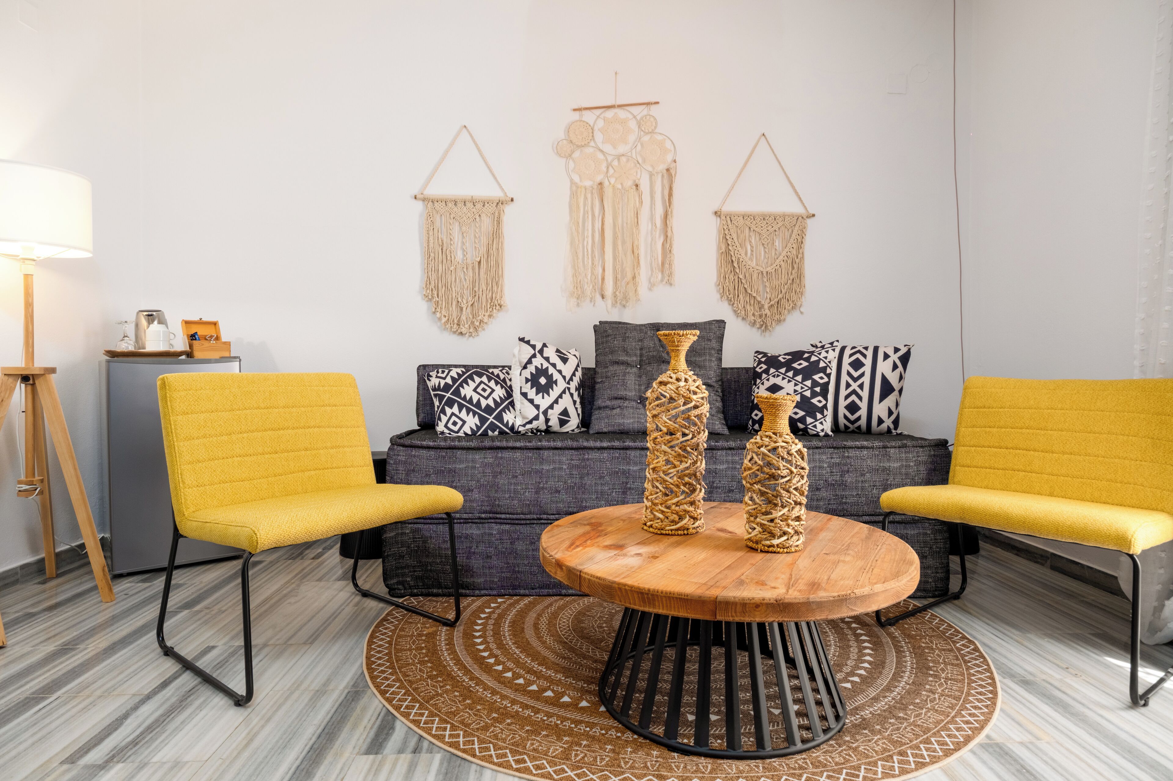 A modern living room with a round wooden table, two yellow chairs, a gray sofa with patterned cushions, and macramé wall hangings.