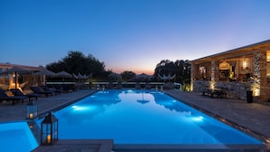 A swimming pool with lounge chairs and umbrellas, surrounded by stone walls and a restaurant with outdoor seating. - Parosland Hotel (Paros)