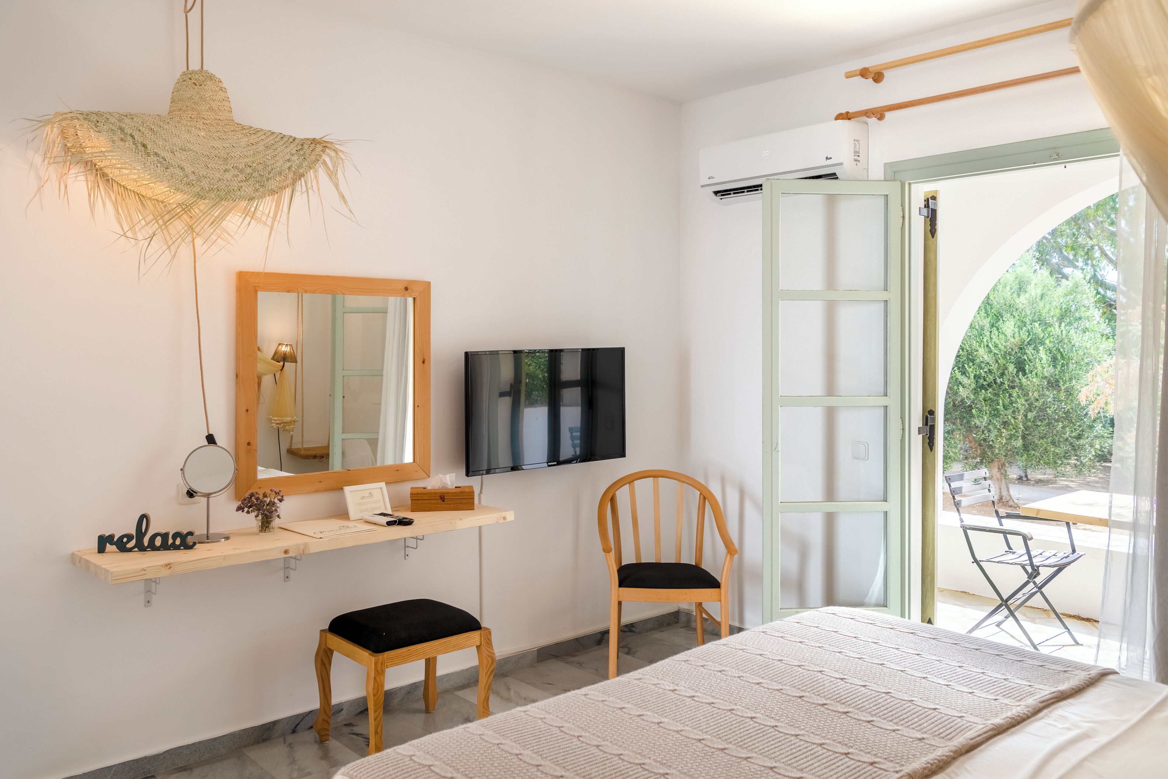 Une chambre à coucher comprenant un lit, une télévision, une chaise, une étagère avec un miroir et une fenêtre avec une porte ouverte donnant sur l’extérieur.