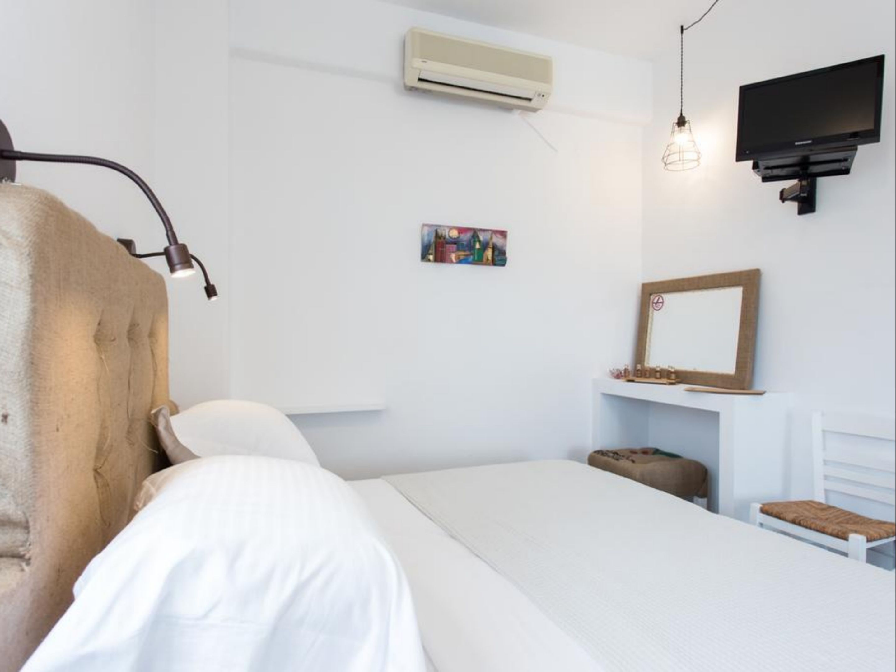 A bedroom with a bed, bedside table, lamp, air conditioning unit, TV, and a small painting on the wall.
