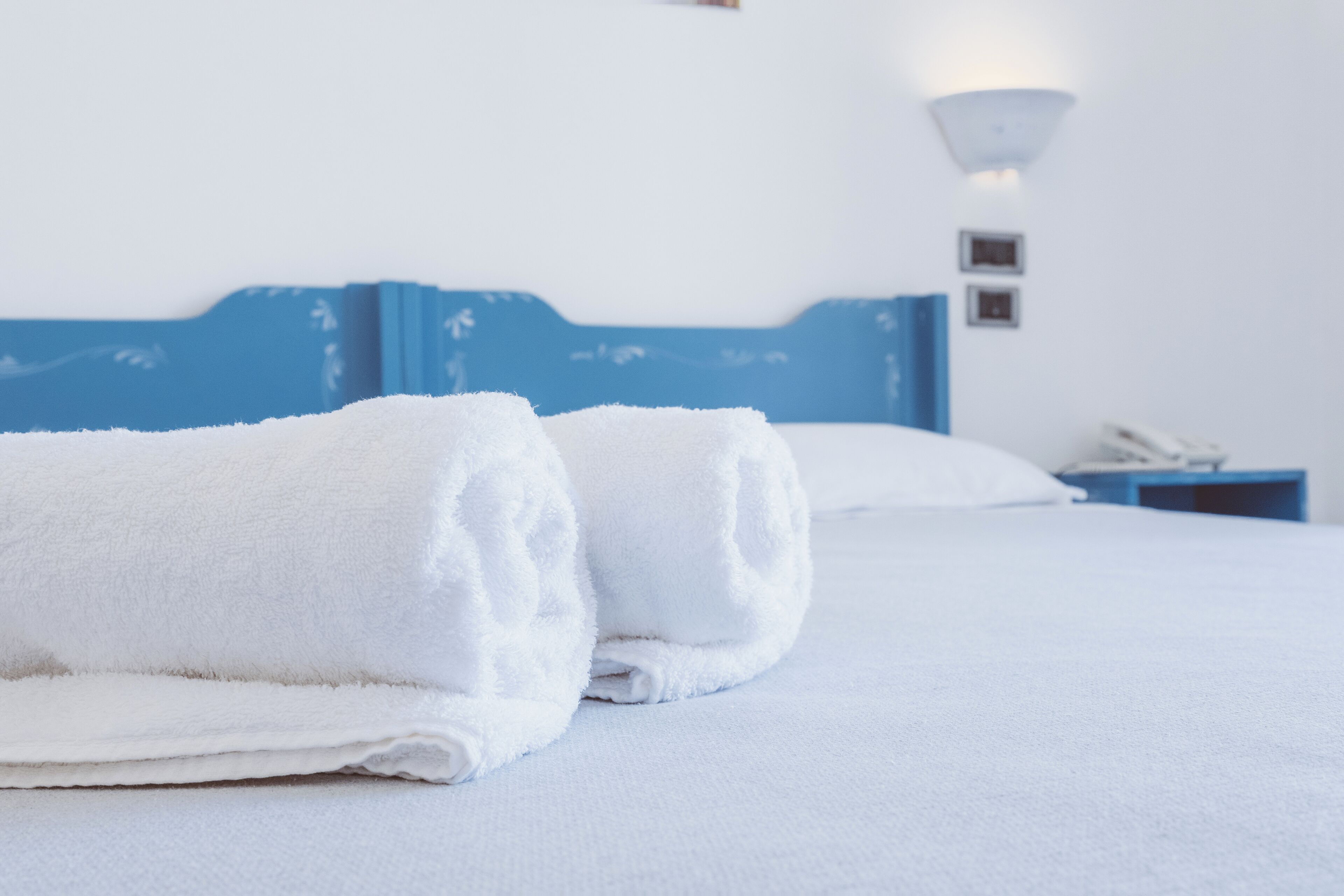 Cama bem arrumada com toalhas brancas, cabeceira azul e mesa de cabeceira com abajur e telefone.