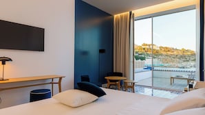 A hotel room with a bed, a desk, a chair, a small table, and a view of a pool and buildings.